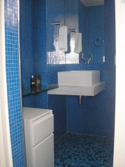 Pequeno sanitário totalmente revestido com pastilhas de vidro azul turqueza. louça branca e pequena bancada de vidro. móvel branco com rodízios completam a decoração simples e utilitária.